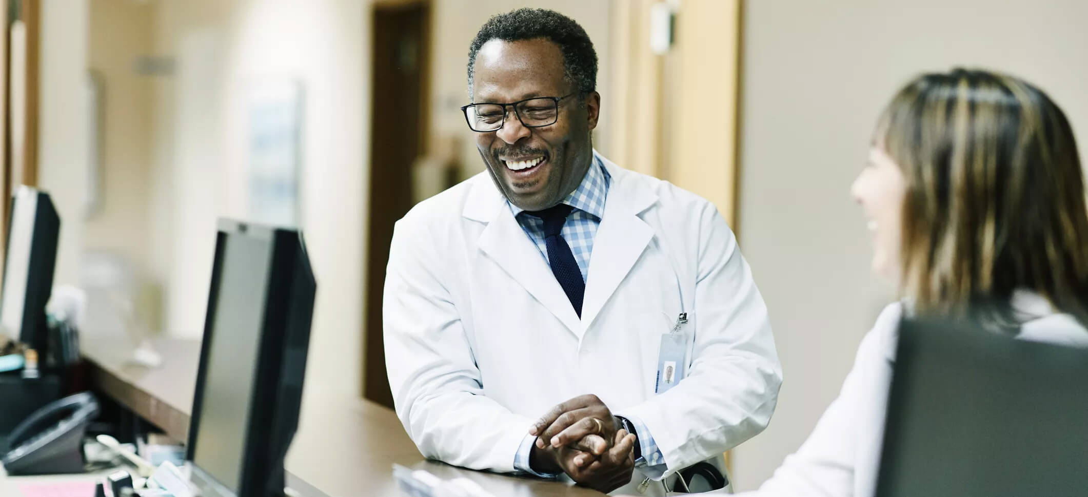 Smiling male doctor in a white coat and glasses talking with a colleague at a hospital workstation, showcasing teamwork and professional interaction in a healthcare setting